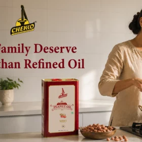 A premium lifestyle kitchen scene featuring a smiling South Asian woman cooking in a bright modern kitchen with white tiled walls. A CHEKKO 5L cold pressed peanut oil tin is placed on the countertop beside raw peanuts, while warm natural sunlight fills the elegant kitchen space, creating a clean, authentic, and healthy cooking atmosphere.